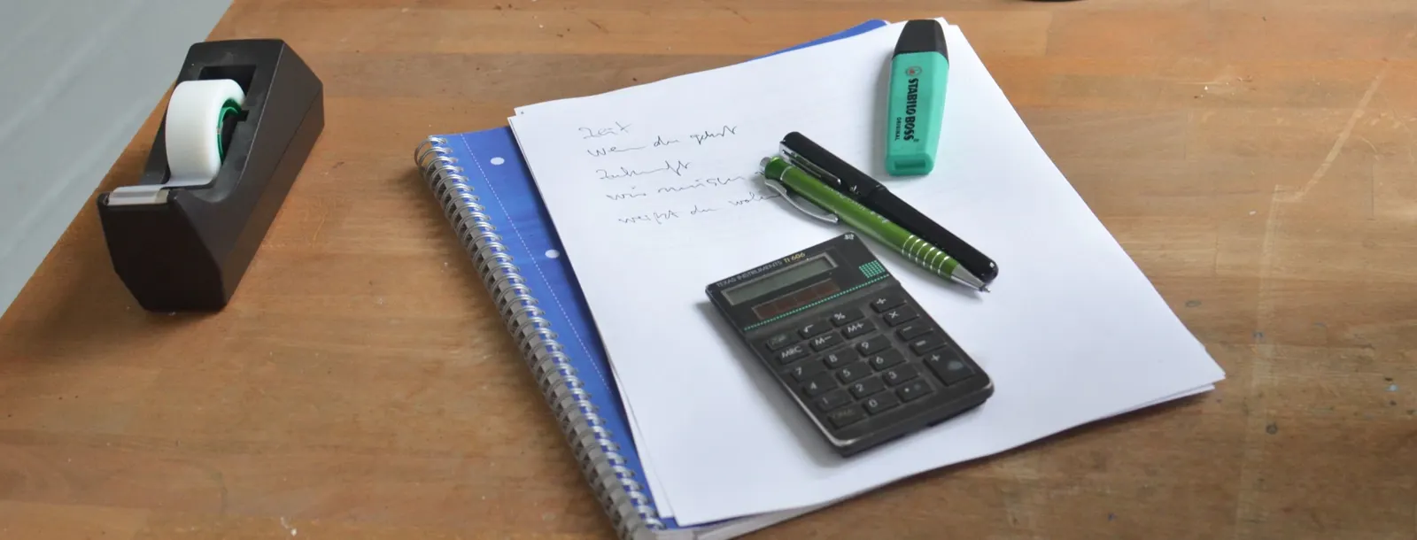 A calculator, a pen and a highlighter on printed pages and a spiral notebook, on a wooden desk