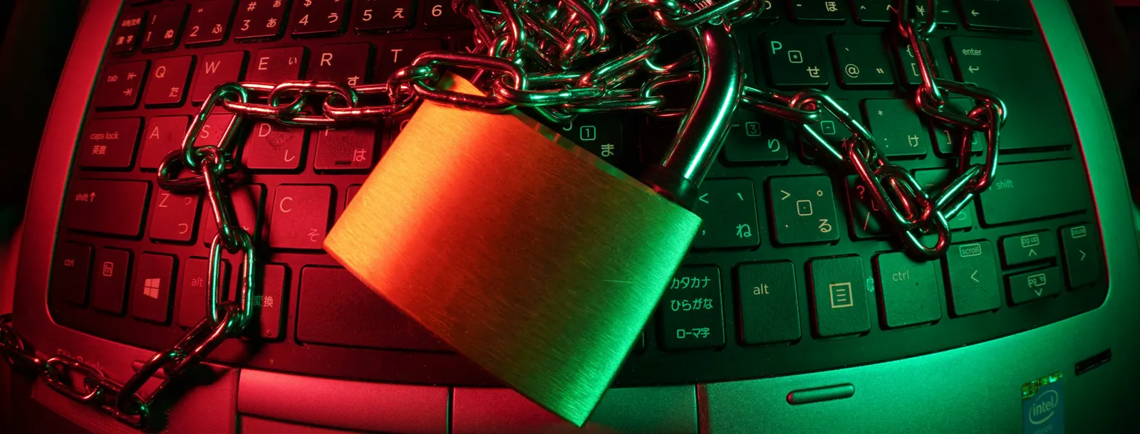 A brass padlock and a steel chain lying on a laptop keyboard under red and green light